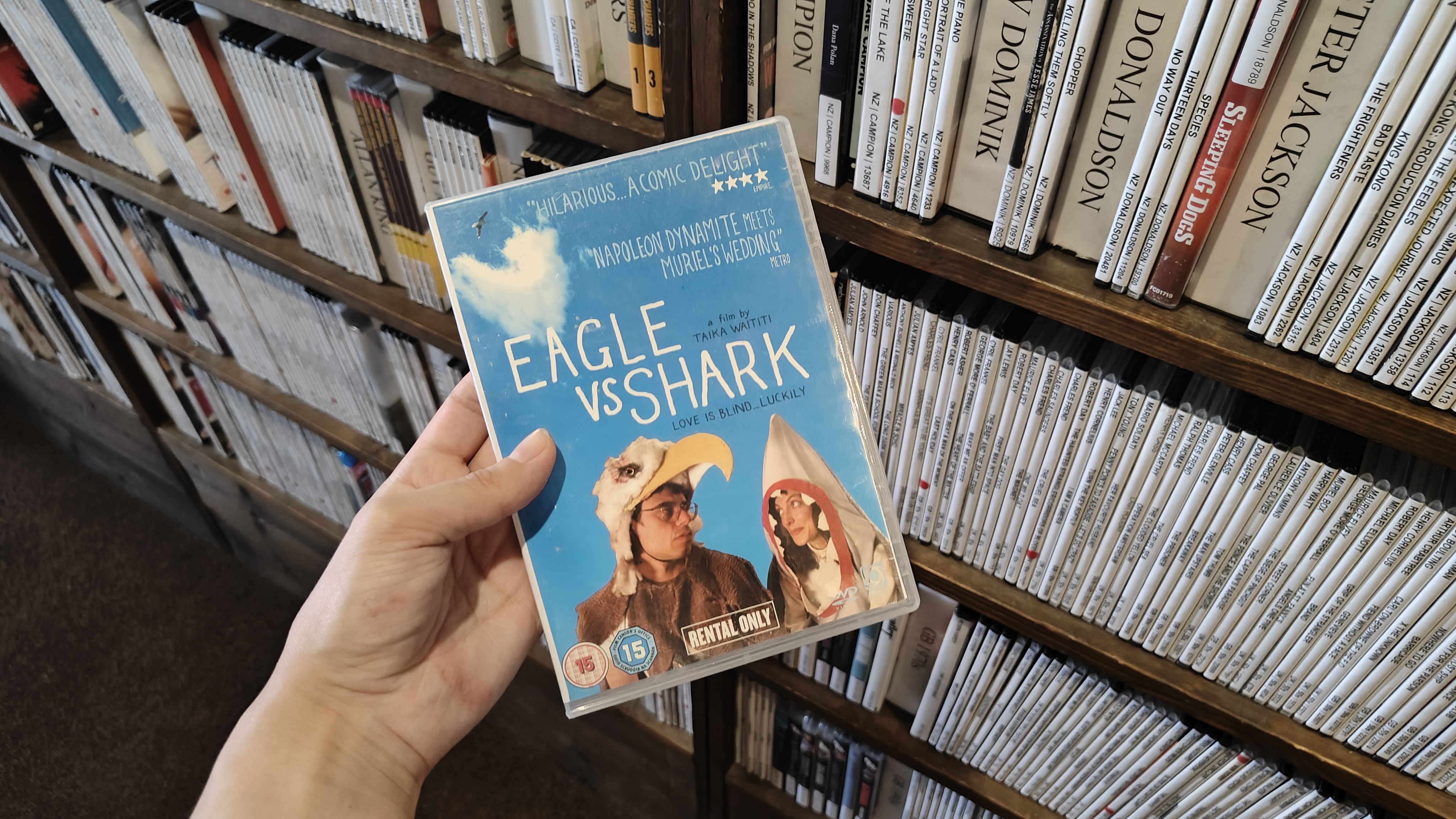 A DVD of 'Eagle vs Shark' is held in front of the camera. It has a blue cover with the two leads dressed in a shark and eagle costume. Behind the DVD are multiple shelves of more DVDs.