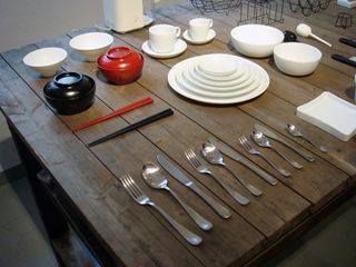 A selection of kitchenware displayed on a table including stainless steel cutlery, white plates, white bowls, white cups, and a red and blue chopstick and food dish bowl.
