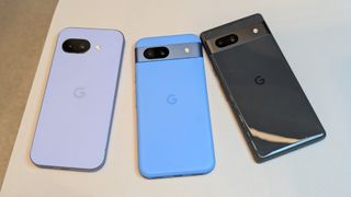 The Iris Google Pixel 9a (left), Bay Google Pixel 8a (center), and Obsidian Google Pixel 7a (right) on a desk, the photo showcasing their different designs.