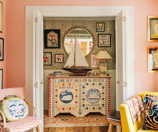 Folk painted side cabinet with patterned wallpaper, mirror, large ship ornament and armchairs