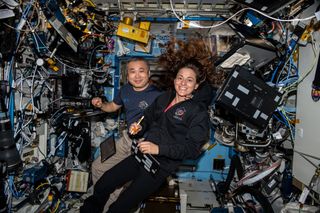two astronauts floating in a space station module crowded with a lot of equipment