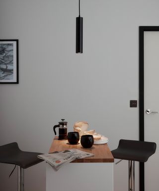 A matt black pendant ceiling light over small kitchen island with newspaper, cafetiere, two coffee cups and sliced bread