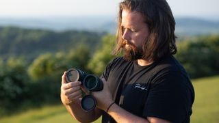 Photo of a man using the Celestron Nature DX 12x56 binoculars against a backdrop of green fields