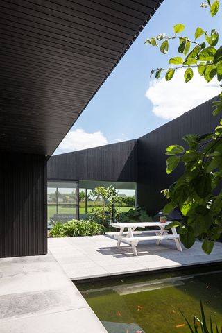 A courtyard in a residential home, with a white bench and table combo on concrete flooring, with a fish pond on one side and green plants on the other side