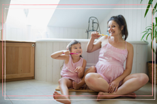 Pregnant mum leaning her teeth with her young daughter - both are sitting on the bathroom floor 