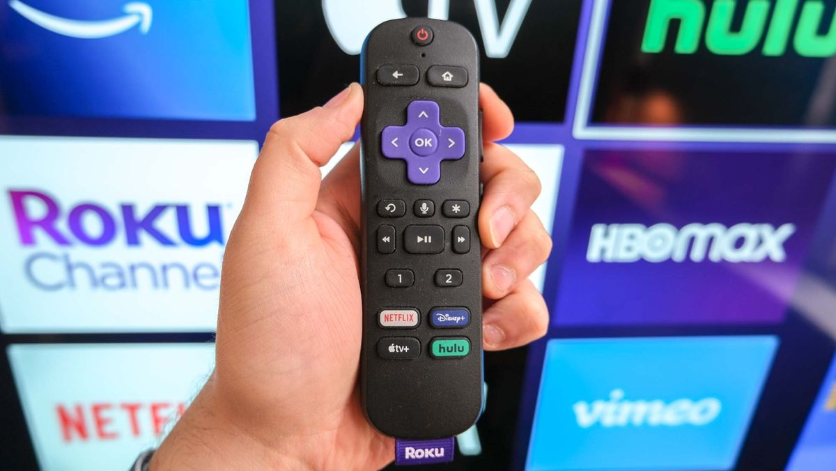 The Roku Voice Remote Pro in hand in front of a TV showing the Roku home screen.