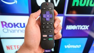 The Roku Voice Remote Pro in hand in front of a TV showing the Roku home screen.