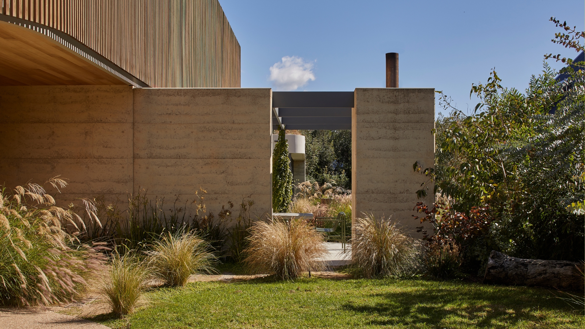 An outdoor garden with wispy grass, a green lawn, tall trees, and an entrance to the backyard and outdoor living room