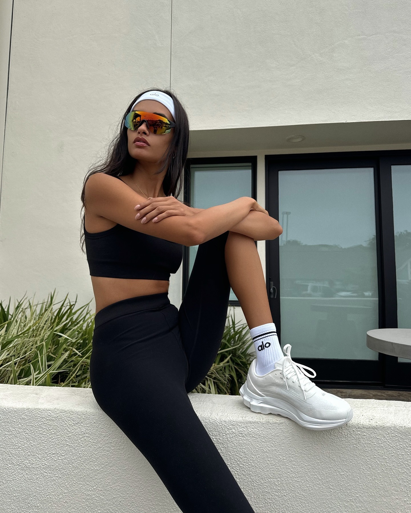 Gizele wears a white headband, black sports bra, black leggings, and white Alo runner sneakers.