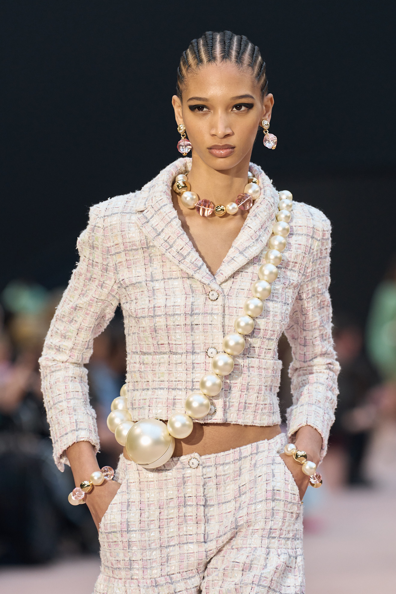 A Chanel model wearing a pink and cream pearl necklace, a pearl over the shoulder bag, and a light pink tweed suit.