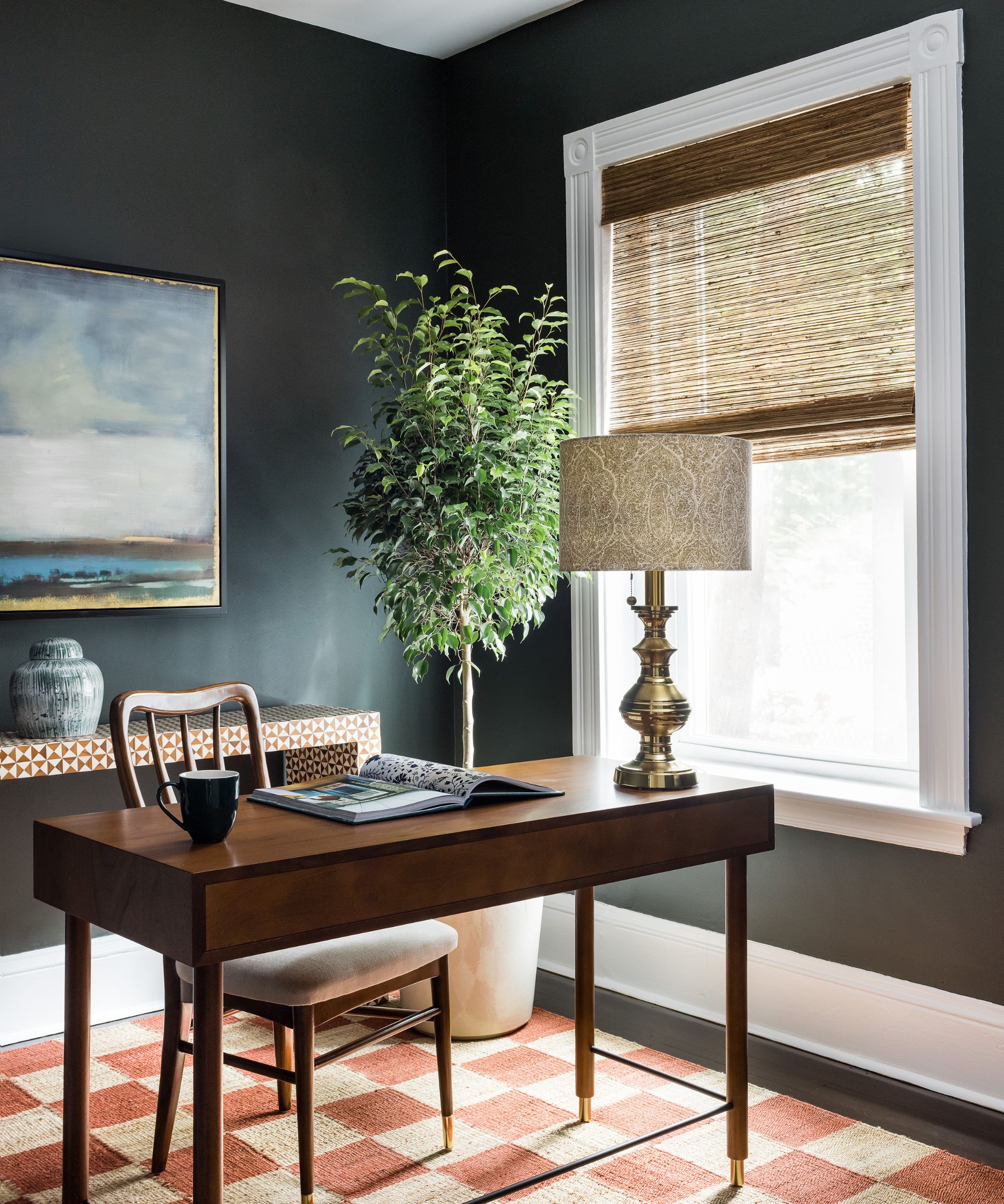 A home office with deep olive green walls, a red and jute checkered rug, white woodwork and window trim, and a wooden desk with a wooden chair, a large hourplant in a pot, and a landscape artwork.