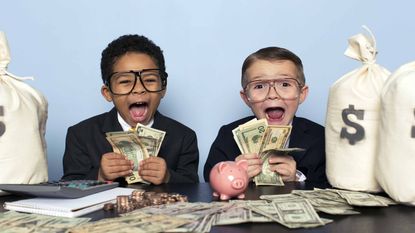 Two cute kids all dressed up like big Wall Street hot shots, with big, excited faces, holding money and sitting at a table with bags of money.