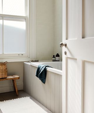 White bathroom with stone colored bath panels, cream metro tiles, toiletries, rustic stool, towel, bathmat