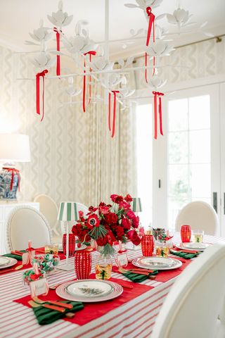 A white dining room with a table set for Christmas dinner. There is a red and white striped table cloth, red placemats, and christmas tree napkins folded on the table.