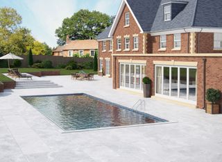 A swimming pool with large form gray rectangular paving outside a brick house.