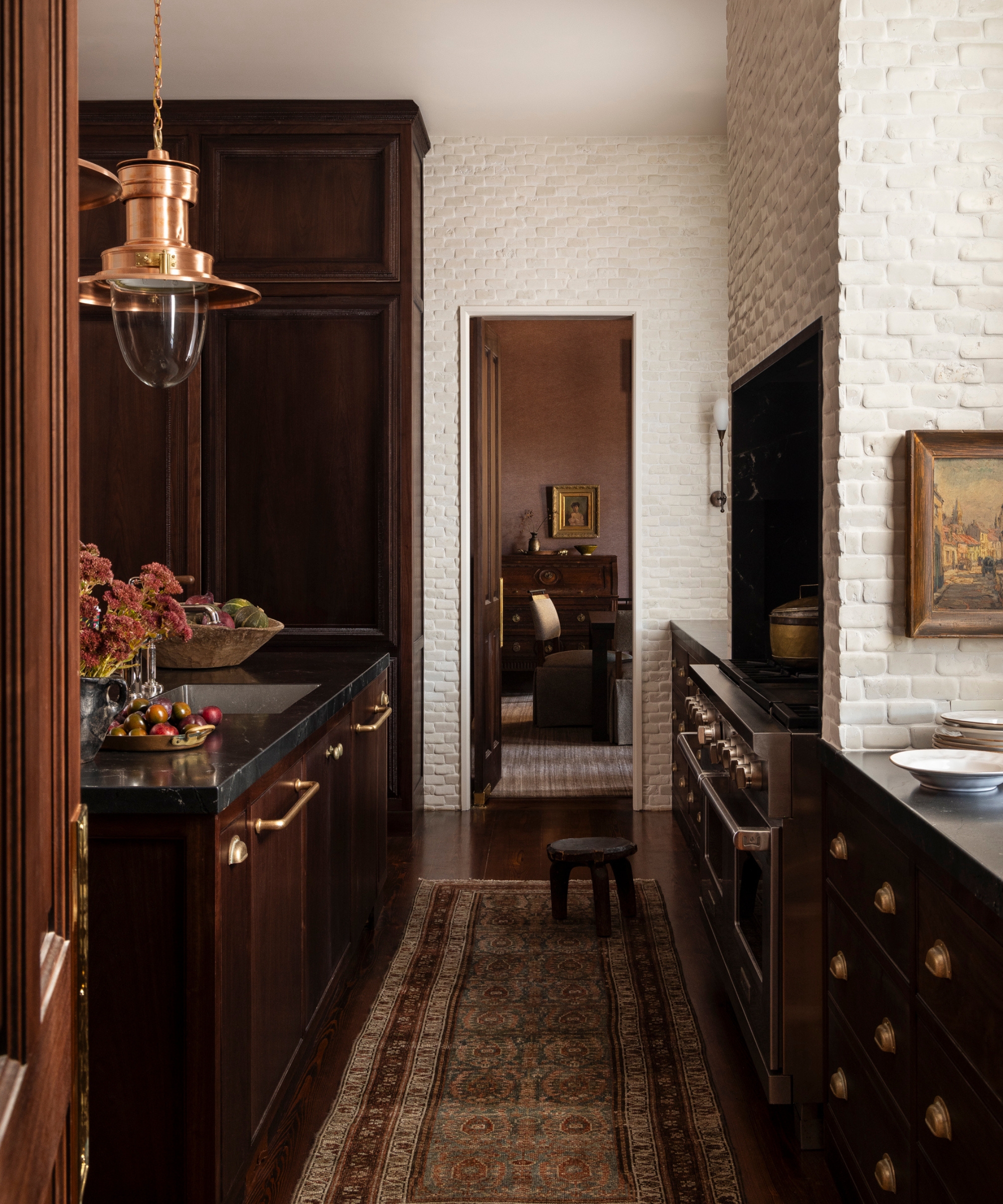A moody kitchen with brown cabinets, black countertops, brass hardware, and a vintage runner