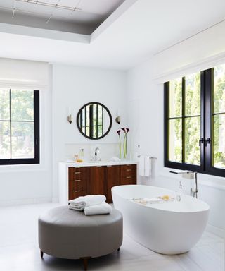 White oval bath, grey ottoman, black framed windows and mirrors, wooden sink