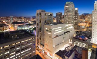 Alternative exterior view of the off-white coloured, irregular shaped San Francisco Museum of Modern Art (SFMoMA) building with the lights on at night. There are multiple buildings surrounding the museum