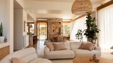 A minimalist living room with off white furniture, beige pillows, sheer window treatments, glass windows, a stone accent wall, potted plants, a wood coffee table, and a woven pendant light