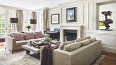 A neutral living room with white walls and ceiling, a large black fireplace, beige sofas with soft pink cushions and a central stone coffee table. Behind are bright, large windows, and two lamps with brown shades.