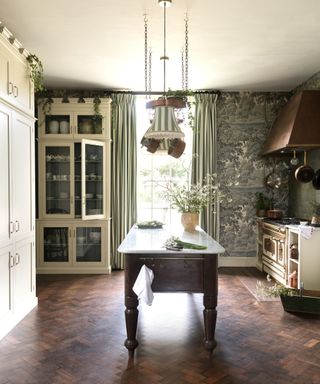 A kitchen decorated with a green mural wallpaper, wooden parquet flooring, and a wooden freestanding island. A traditional range cooker sits in the corner in a cream and brass colorway and a ages brass range hood, white the cabinets have been painted in a matching cream color and topped with castellations at the top