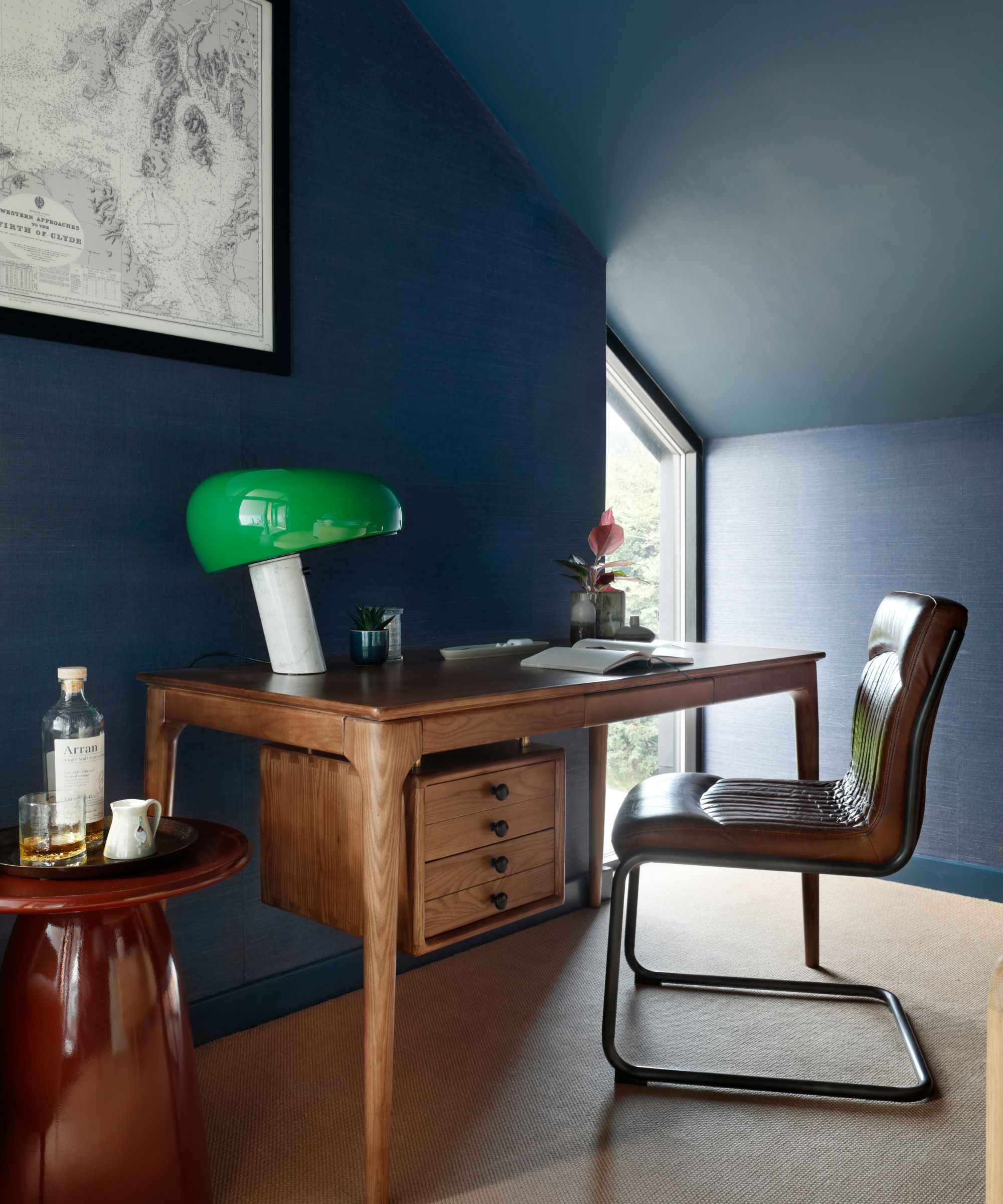 Home office with sloped ceiling painted dark blue, grasscloth dark blue walls, midcentury wooden desk with midcentury leather desk chair, red lacquer side table and modern glass green table lamp