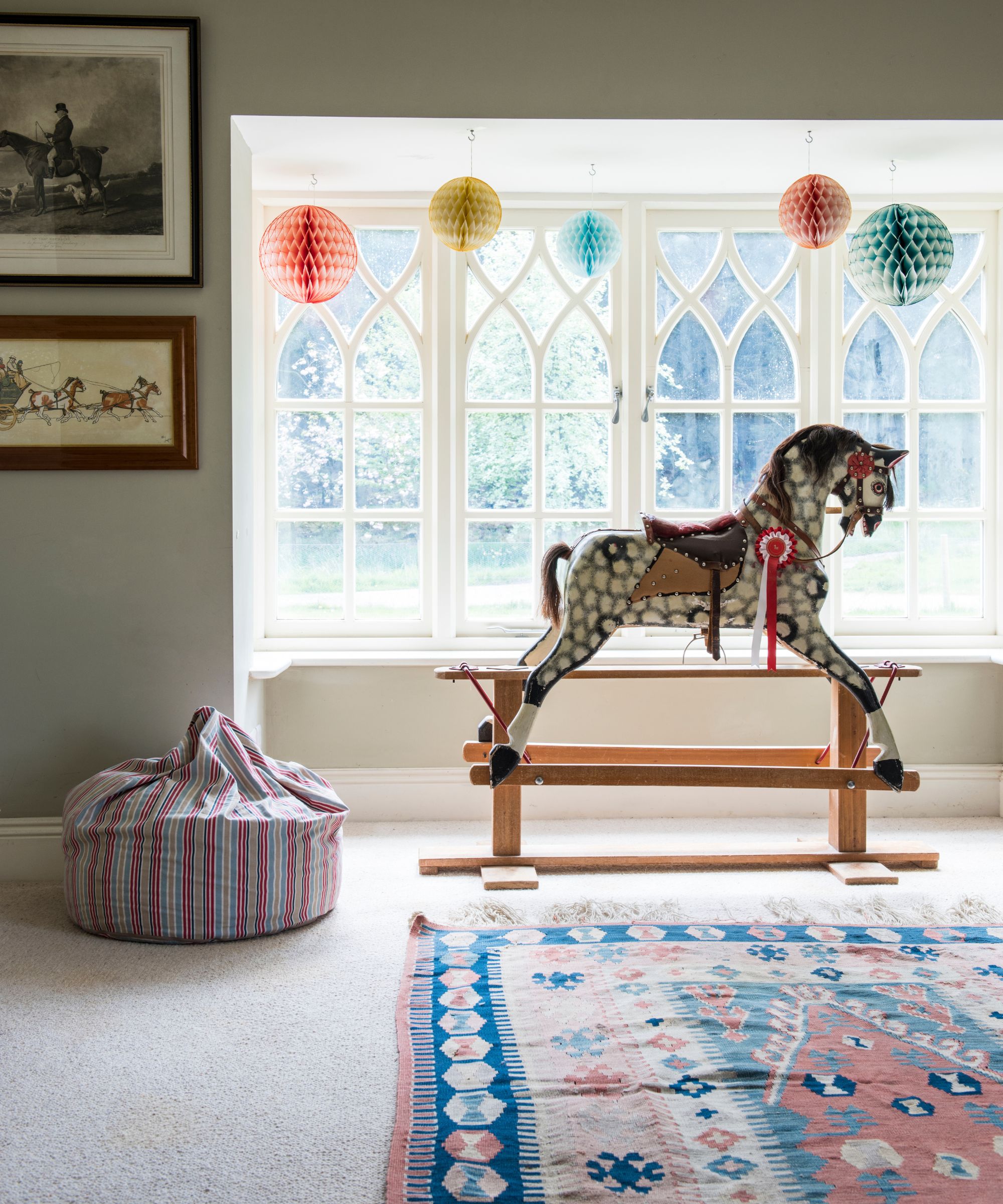 Playroom with a patterned rug, rocking horse in front of a large window, striped beanbag, and artworks of horses.