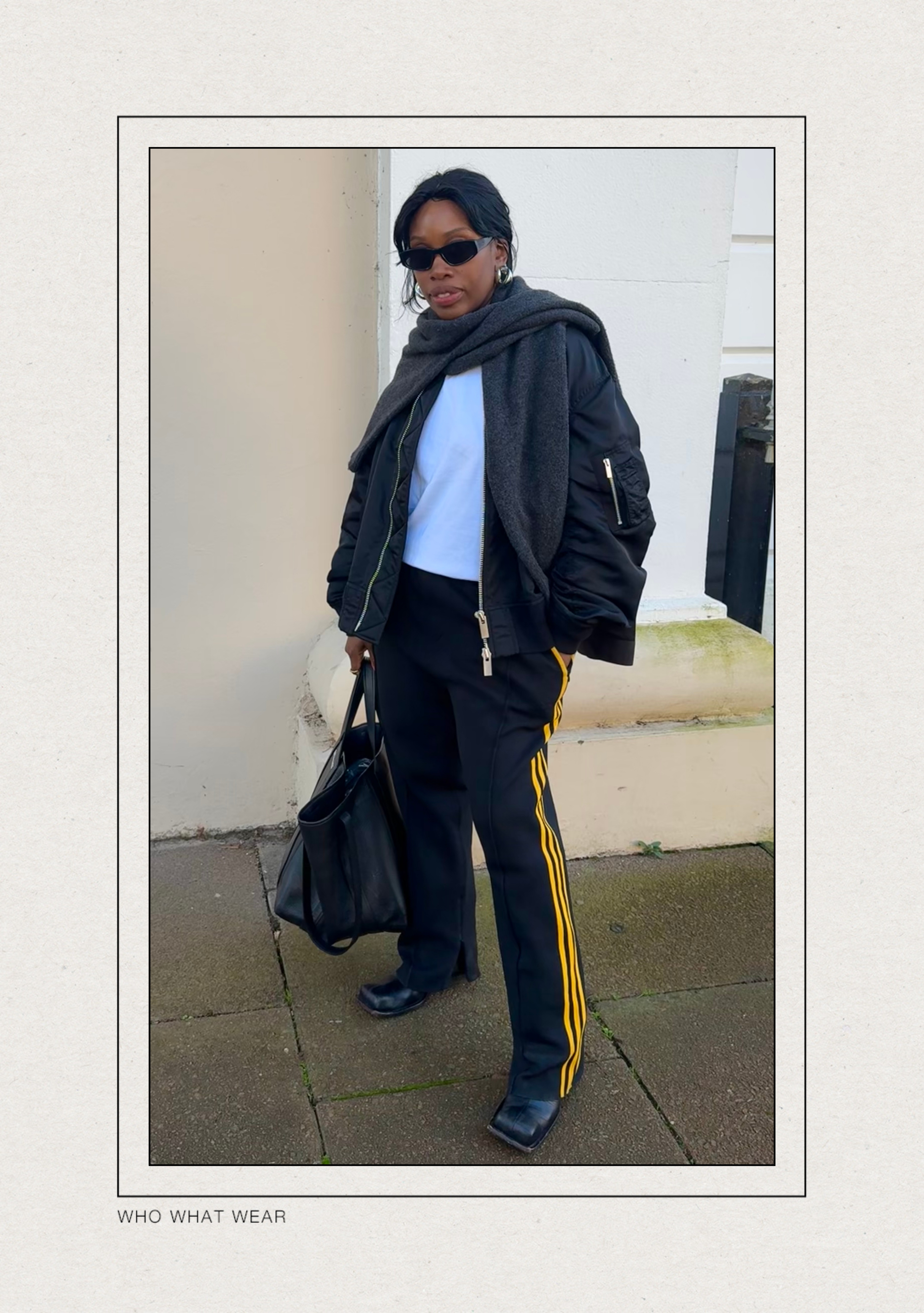 @anuogugbesan wears a black bomber jacket with a white t-shirt, Adidas joggers with yellow side stripes and black boots whilst standing in the street