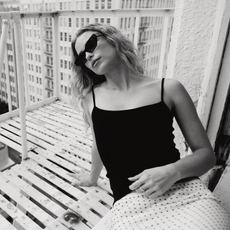 Elle Chapman leans out her window onto her balcony in NYC wearing a black tank top and white skirt with black polka dots. She is wearing black sunglasses. 