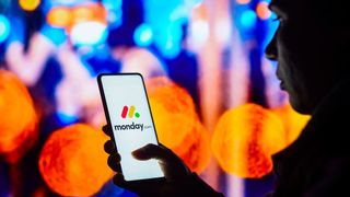 A person holding a smartphone with the logo for monday.com displayed. The person is silhouetted by a background of bright and colourful street lights