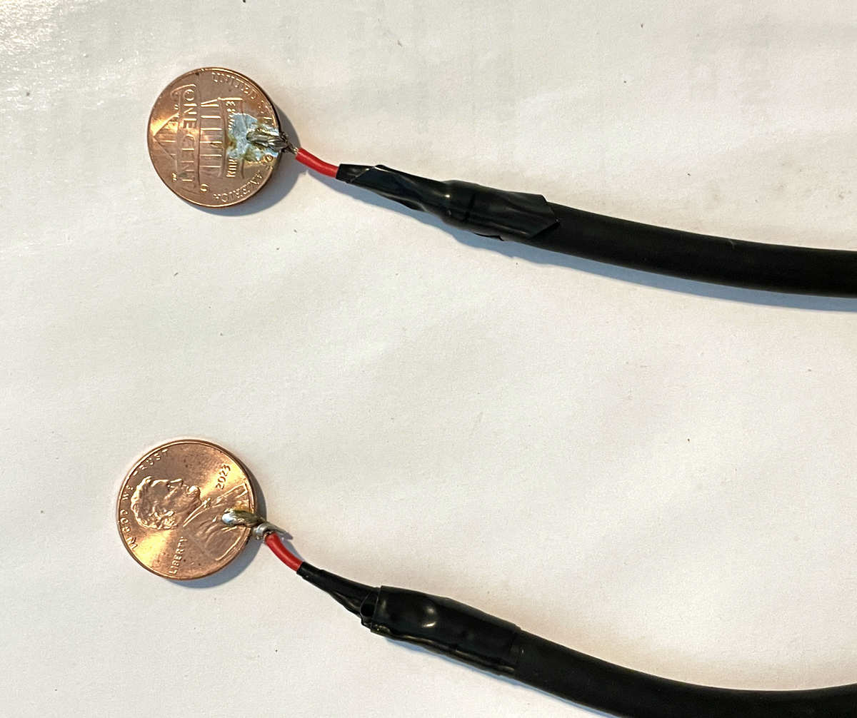 A top-down, close-up view of two handmade audio leads consisting of black insulated cables soldered directly to copper US one-cent coins.