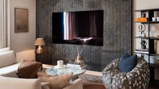 TV mounted onto panelled wall in small living room with patterned armchair, white L-shaped sofa, lamps, round marble coffee table, and shelving on the walls