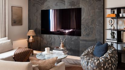 TV mounted onto panelled wall in small living room with patterned armchair, white L-shaped sofa, lamps, round marble coffee table, and shelving on the walls