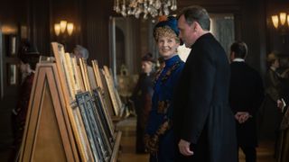 Cynthia Nixon in a blue dress and hat as Ada stands with Robert Sean Leonard in a black suit as Rev Forte admiring a picture in The Gilded Age 2.