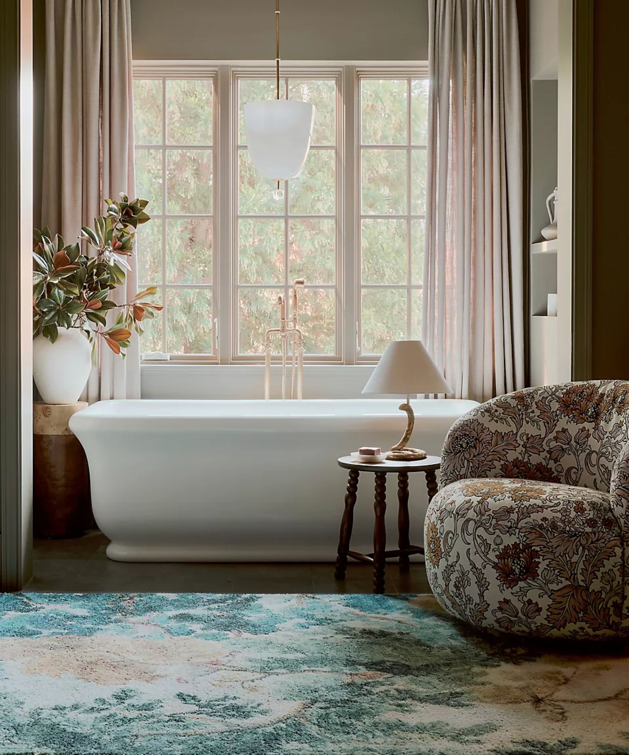 moody pink bathroom with a large window and a freestanding bath styled with a wooden bobbin stool and lamp, tapestry print upholstered armchair and a floral mural style rug