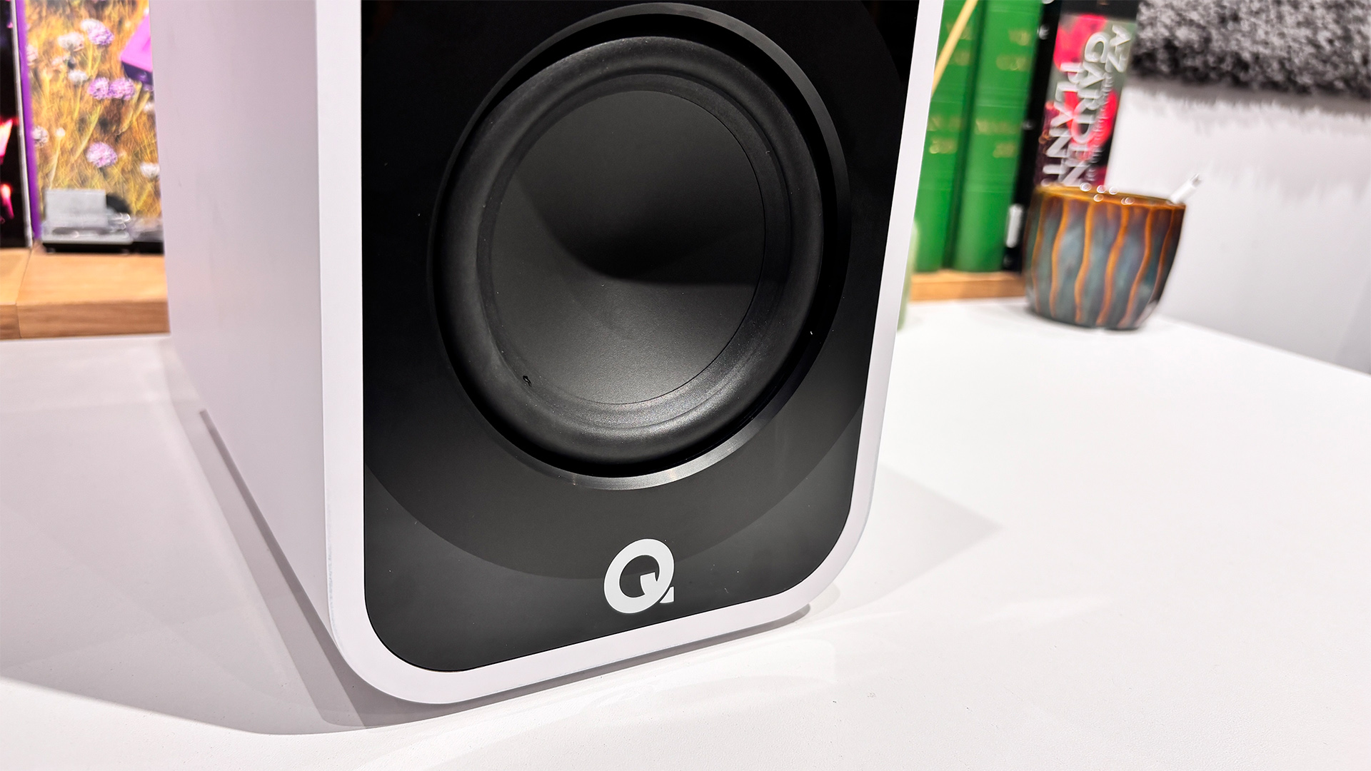 Q Acoustics 5010 standmount speakers on white surface in front of busy bookshelf showing bottom of speaker with mid/bass unit and Q Acoustics logo