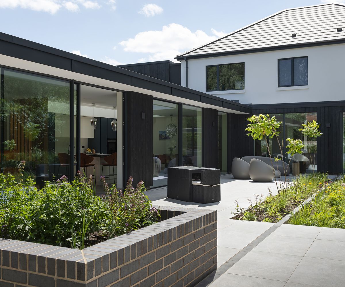 A contemporary extension clad in dark timber and large glazing with a pale grey patio in front with modern garden furniture