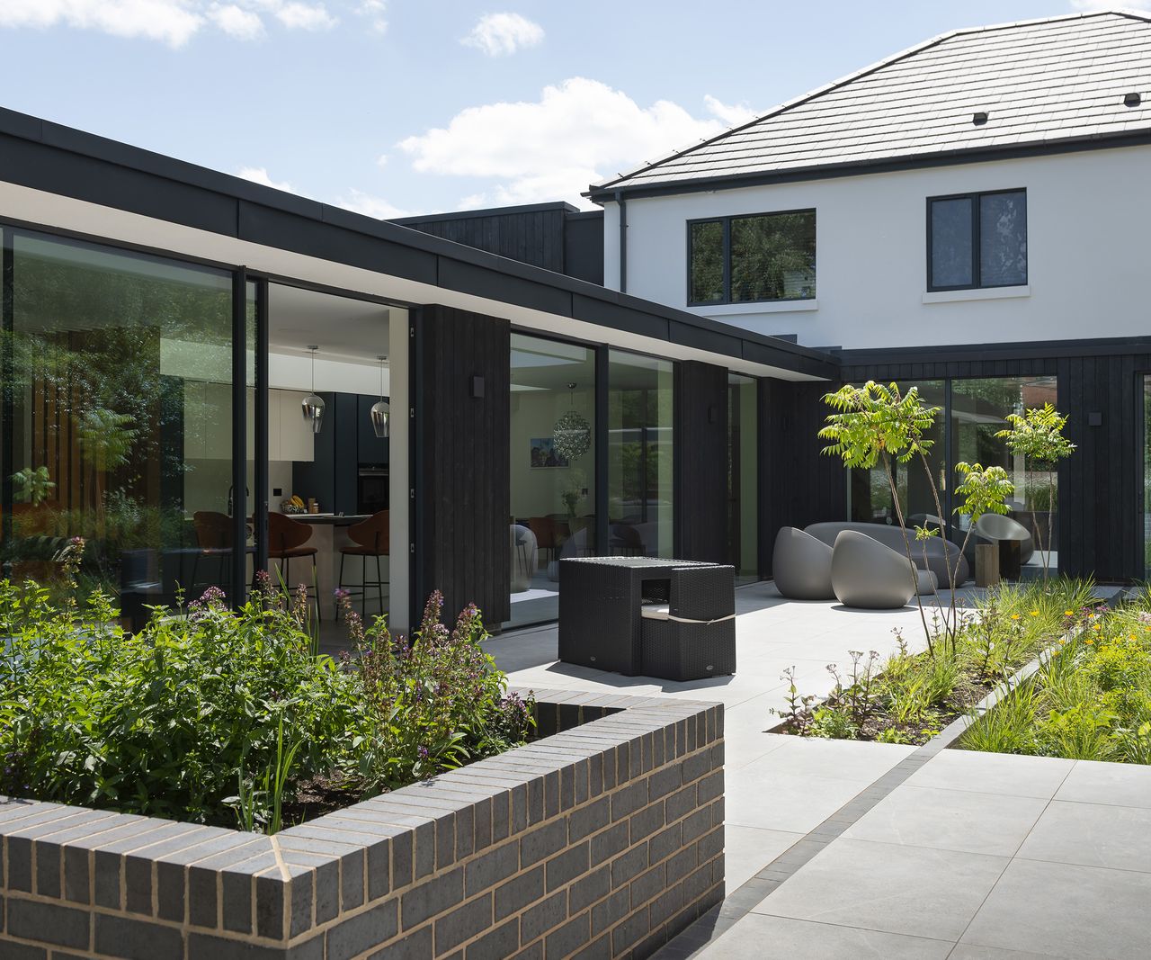 A contemporary extension clad in dark timber and large glazing with a pale grey patio in front with modern garden furniture