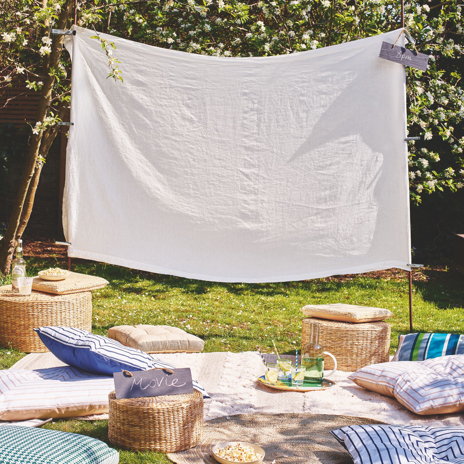 garden with white sheet for projector/cinema, cushions, rugs, blankets and sign saying 'movie', rattan side table and footstools