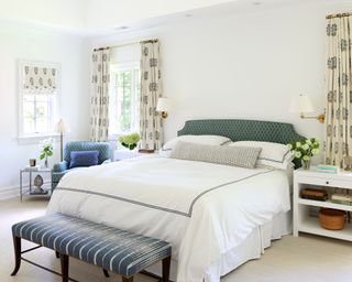 Master bedroom with patterned curtains and matching blind, large bed with white bedding, blue patterned headboard, bench at end of bed, cream carpet, white painted walls, two bedside tables, chair in corner with matching upholstery to headboard
