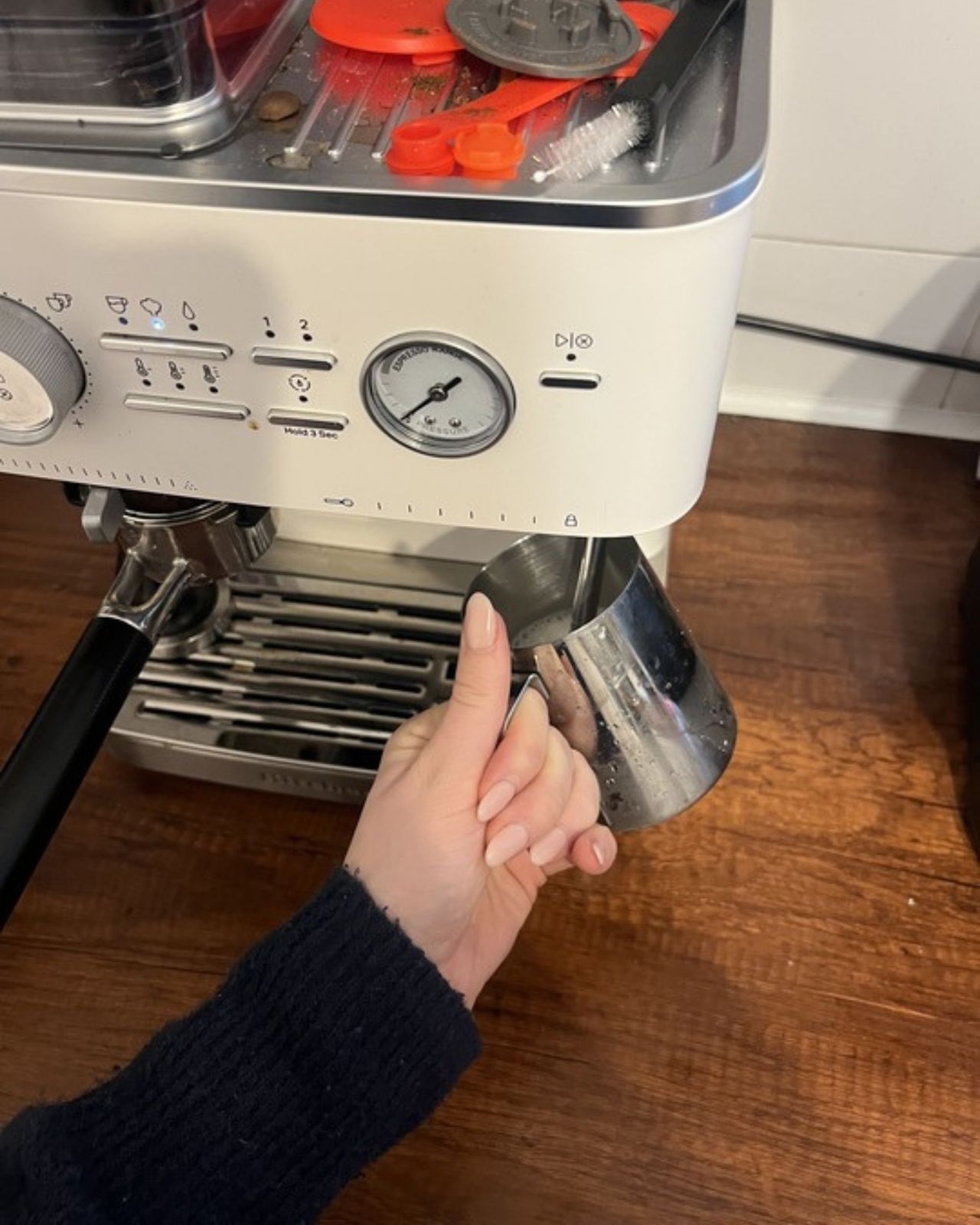 Image of a hand holding a milk jug under a steaming wand attached to a white espresso machine.