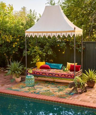 A backyard space with lemon trees, a white tent with a colorful bench and red, yellow, and blue throw pillows underneath, a tropical rug underneath this and plants next to it, and a swimming pool
