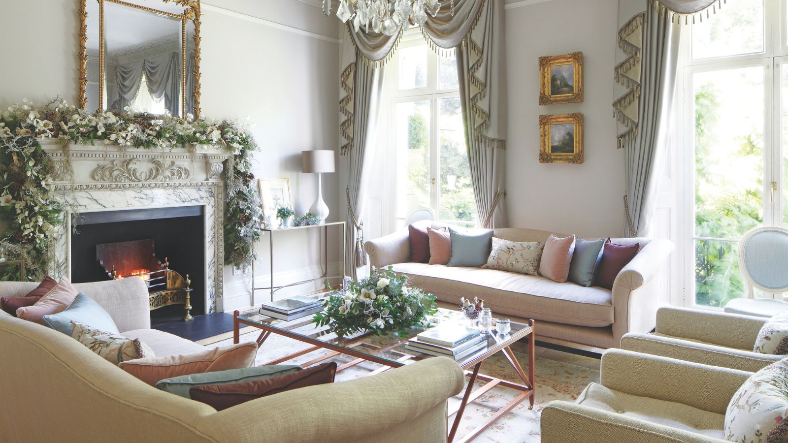 An elegant living room with white walls, white and gold fringe curtains over large windows to the back of the room, and a large marble mantelpiece to the left with a lit fire, large flower garland, and large gold mirror above. In the center of the room is a large glass coffee table with flowers, and upholstered pink and yellow couches and chairs surrounding it with mismatched colored cushions. Beneath the coffee table is a patterned rug.