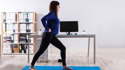 Woman in business attire and bare feet stands in a staggered stance on an exercise mat, in front of an office desk with a monitor on it