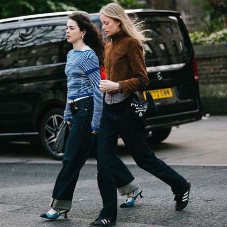 Two woman walk across the street. One is wearing a longsleeve blue striped t-shirt, cuffed jeans, and heels with socks. The other is wearing a brown suede jacket, black jeans, and Adidas sambas.