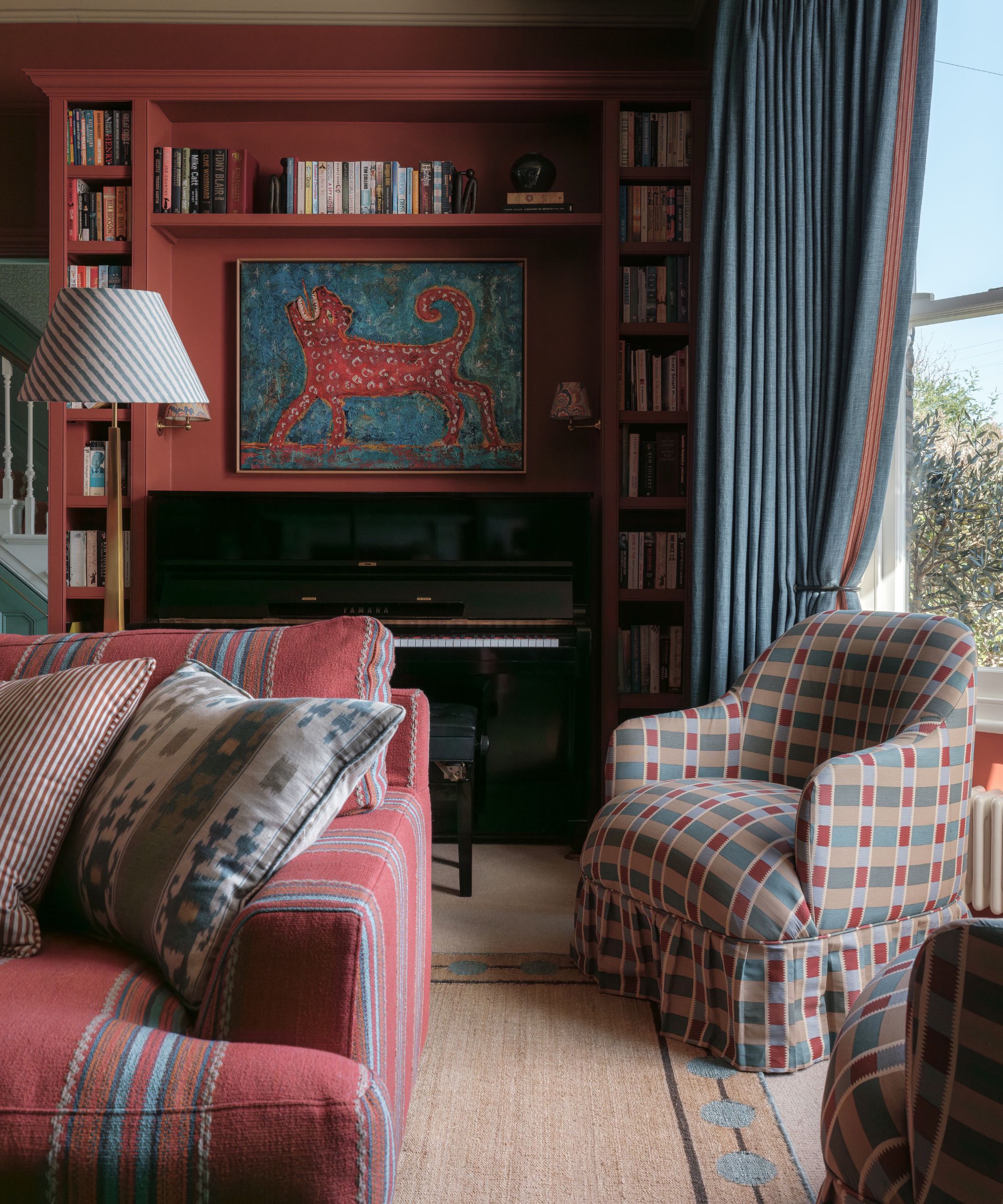 A cozy maximalist living room with dark red built-ins, a bay window with muted blue curtains, a red striped sofa with patterned cushions, checkered armchair, and a jute rug.