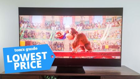 The TCL QM7K resting on top of a media console while it displays a scene from "The Super Mario Bros. Movie." A badge superimposed onto the bottom-left corner reads: "Tom's Guide Lowest Price".