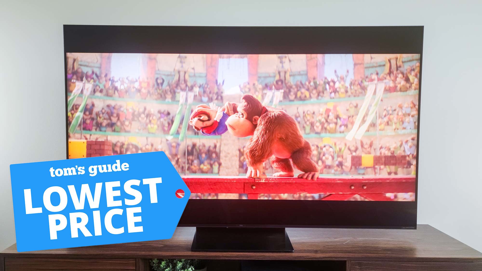The TCL QM7K resting on top of a media console while it displays a scene from "The Super Mario Bros. Movie." A badge superimposed onto the bottom-left corner reads: "Tom's Guide Lowest Price".