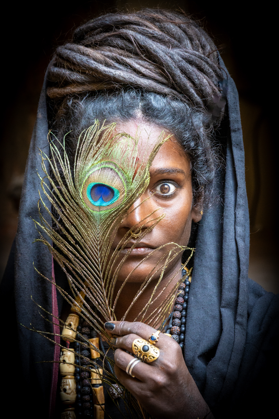 A person wearing a grey cloth head wrap holds a vivid peacock feather across half their face, revealing one intense eye, adorned with layered beaded necklaces and ornate rings.