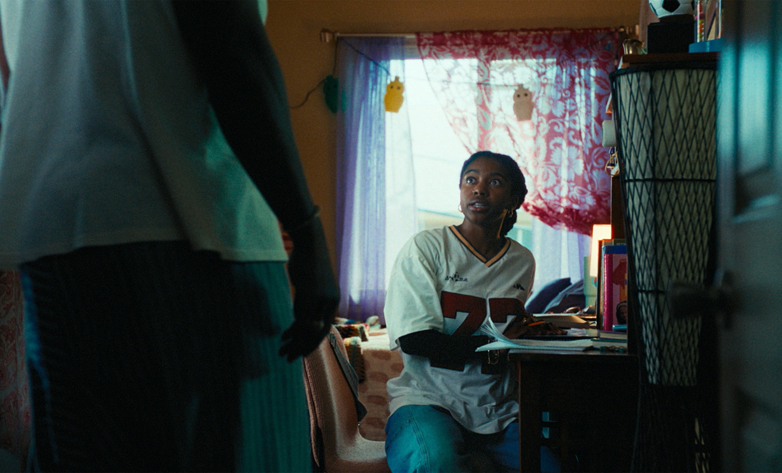 Adia as Fatiha sits at a bedroom's desk in front of a large window draped with red lace, with an unseen person standing in front of her, in a still from 'Your Mother Your Mother Your Mother.'